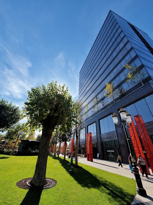 A modern glass office building stands tall under a clear blue sky, reflecting light and partial clouds. In the foreground, a well-maintained lawn with neatly trimmed trees and red pillars lines a path where a few people are strolling. A vintage-style lamppost adds a touch of elegance to the scene.