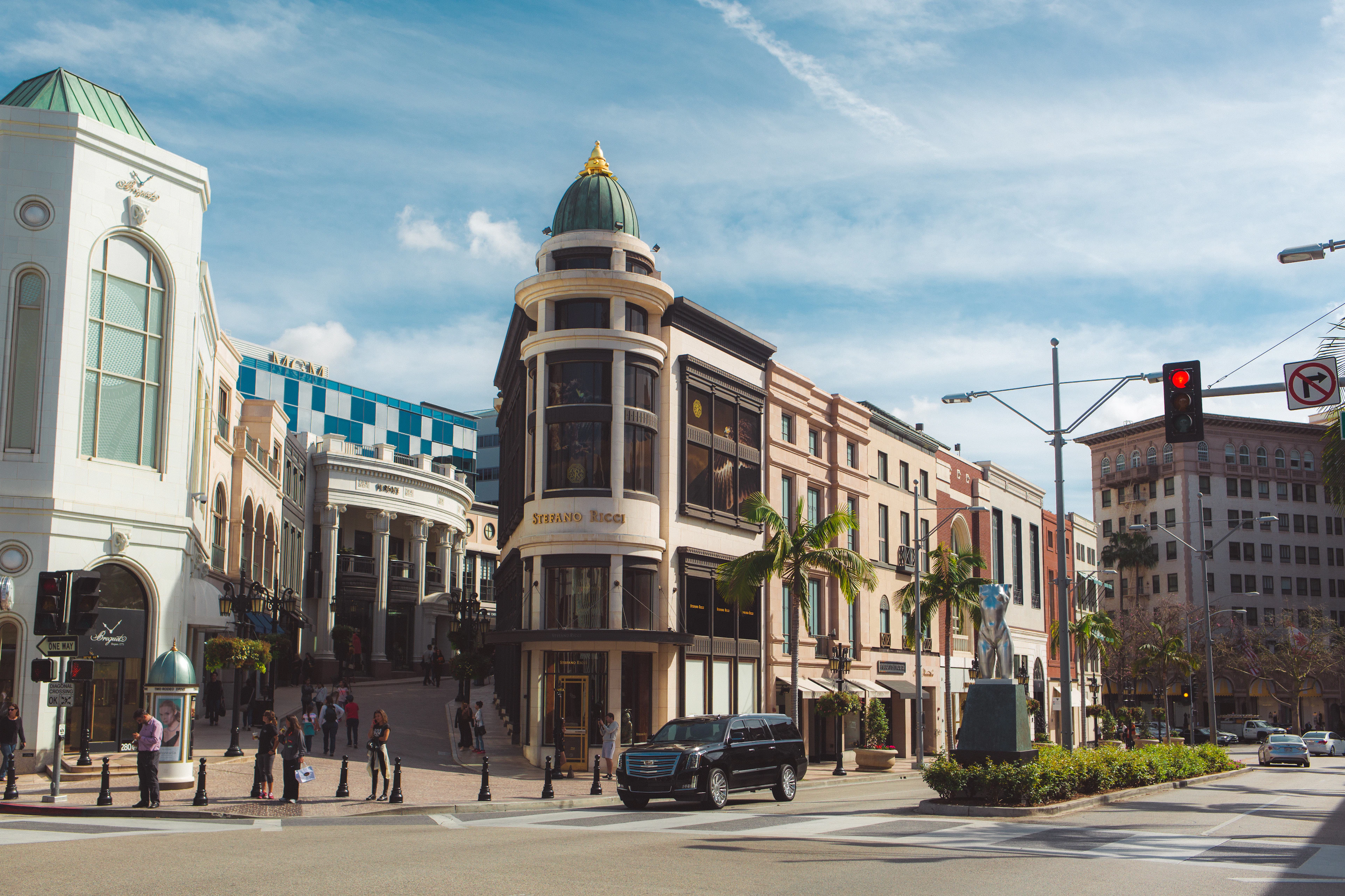 The corner of Rodeo Drive in Beverly Hills on a bright day, a copper-domed turret above the boutique frontages, palms and a bronze statue along the sidewalk and a black SUV waiting at the crossing.