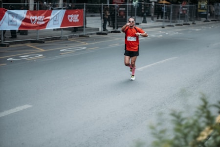 A person wearing a bright orange shirt and dark shorts is running on a city street. The runner appears to be in a race or marathon setting, indicated by the presence of a number bib on their shirt. There is a red and white banner in the background with writing on it, and the street is marked with route numbers. Metal barriers line the street, suggesting an organized event.