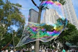 A giant, iridescent bubble floats in a park setting surrounded by tall buildings and lush green trees. A person holds the bubble wand to the left, creating a mesmerizing display of colors in the sunlight. Numerous people are gathered in the background, enjoying the outdoor scene.