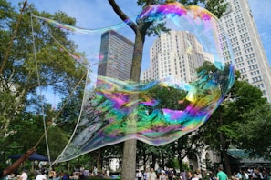 A giant, iridescent bubble floats in a park setting surrounded by tall buildings and lush green trees. A person holds the bubble wand to the left, creating a mesmerizing display of colors in the sunlight. Numerous people are gathered in the background, enjoying the outdoor scene.