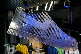 A large, artistic installation resembling a sneaker made of translucent material hangs from the ceiling, illuminated by spotlights with blue tints. Below, colorful clothing items such as yellow, green, and blue jackets are displayed on racks in a retail store. The setting includes a dark, industrial-style ceiling with visible lighting fixtures.