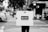 A person stands on a city street holding a large rectangular sign that reads 'BLICK' with a website address below it. The scene includes cars in the background, a crosswalk, trees, and buildings along the sidewalk, all captured in black and white.