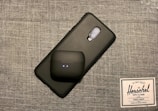 A smartphone with a textured black case is placed on a gray woven fabric surface. On top of the phone, there is a black wireless earbud case with two small indicator lights. Beside these items, there is a label or tag from Herschel Supply Co., featuring their logo and branding text.