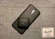 A smartphone with a textured black case is placed on a gray woven fabric surface. On top of the phone, there is a black wireless earbud case with two small indicator lights. Beside these items, there is a label or tag from Herschel Supply Co., featuring their logo and branding text.