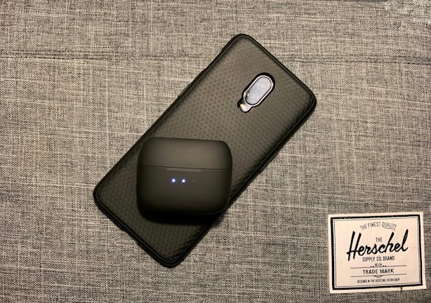 A smartphone with a textured black case is placed on a gray woven fabric surface. On top of the phone, there is a black wireless earbud case with two small indicator lights. Beside these items, there is a label or tag from Herschel Supply Co., featuring their logo and branding text.