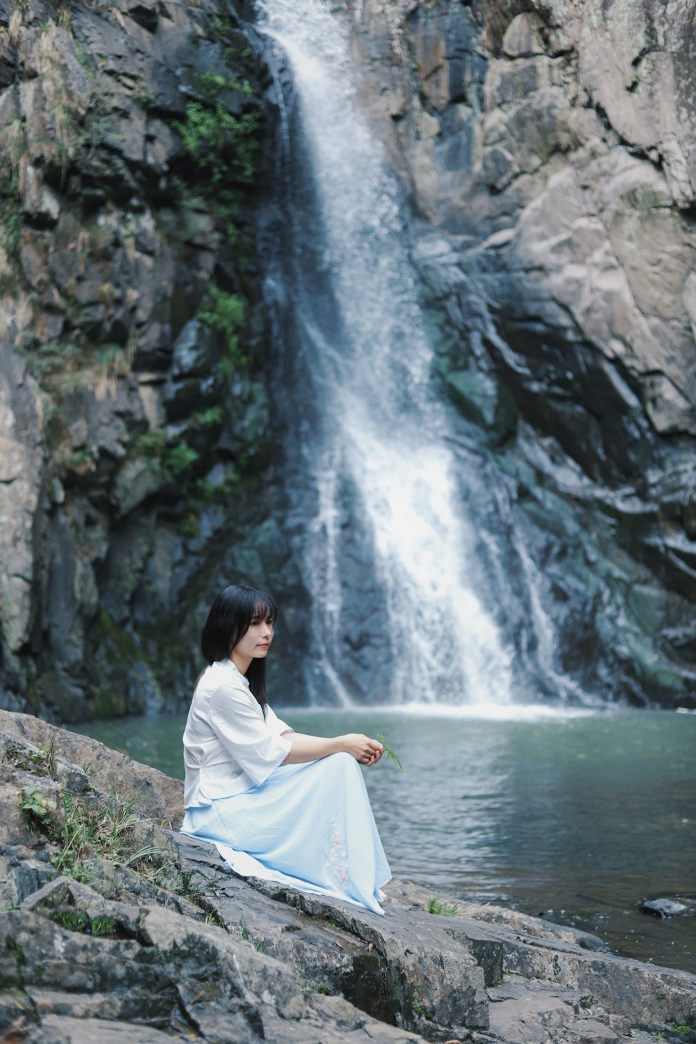 Frau im weißen Kleid sitzt auf einem Felsen neben Wasserfällen
