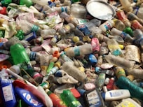A pile of discarded plastic bottles and containers, some with colorful labels, mixed with other waste and debris. The items are scattered haphazardly, indicating pollution and environmental neglect.