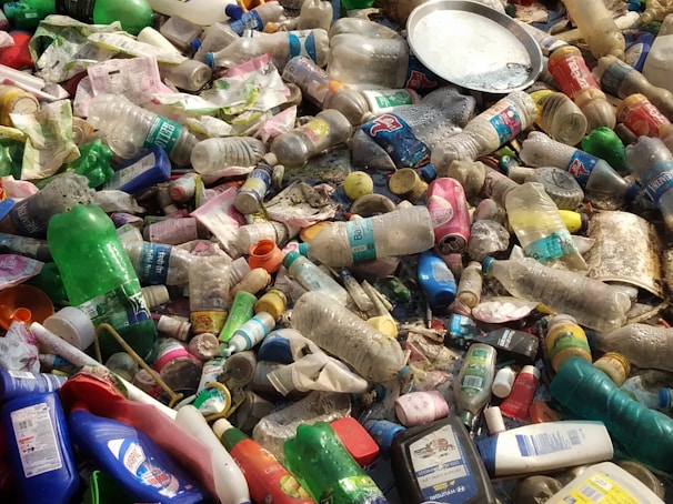 A pile of discarded plastic bottles and containers, some with colorful labels, mixed with other waste and debris. The items are scattered haphazardly, indicating pollution and environmental neglect.