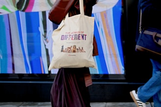 A person is walking while carrying a canvas tote bag with a design promoting diversity, reading 'The Power of Different' along with images of various dogs. The background features a vibrant, abstract display with vertical streaks of color.