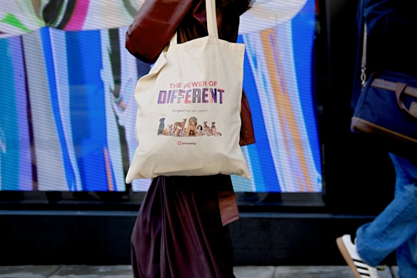 A person is walking while carrying a canvas tote bag with a design promoting diversity, reading 'The Power of Different' along with images of various dogs. The background features a vibrant, abstract display with vertical streaks of color.