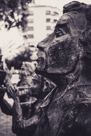 A close-up view of a weathered bronze sculpture depicting a human profile in an outdoor setting. The texture of the sculpture is rough and intricate, capturing the facial features in a stylized manner. In the background, blurred outlines of trees and a building can be seen.