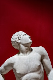 A classical marble statue depicting a male figure with curly hair, looking upwards with a relaxed posture against a striking red background. The sculpture is characterized by smooth textures and defined musculature.