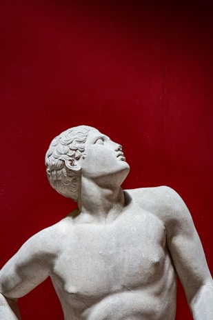 A classical marble statue depicting a male figure with curly hair, looking upwards with a relaxed posture against a striking red background. The sculpture is characterized by smooth textures and defined musculature.