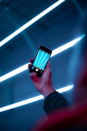 A person holds a smartphone with a dark screen displaying bright blue lines. The background features a series of parallel neon lights creating a futuristic ambiance. The overall setting appears to be dark with these luminous elements standing out.