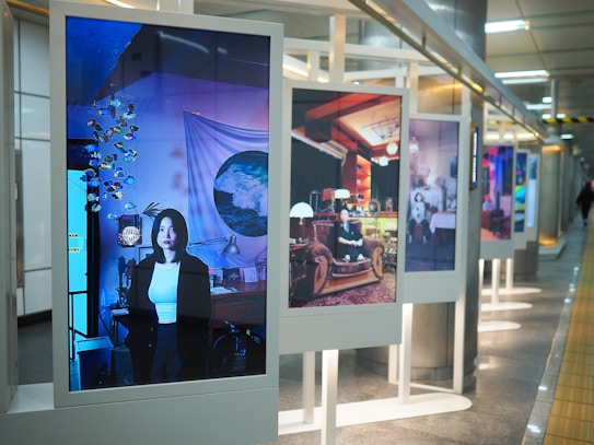 A series of large digital displays are positioned in a subway or transit station setting, each showing different artistic portraits. The nearest display features an image of a woman in a room with blue lighting and decorative items like a mobile and wall art. The scene is modern and artistic, with a focus on individual expressions and creativity.