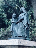 A bronze statue depicts a woman in a hooded robe sitting and reading a book with a young girl standing beside her. The figures are positioned on a stone pedestal surrounded by lush green foliage.