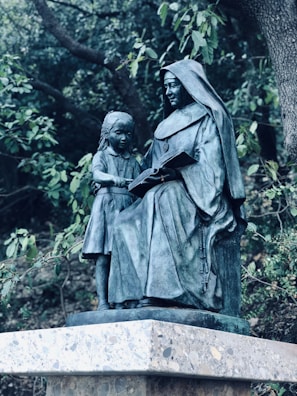 A bronze statue depicts a woman in a hooded robe sitting and reading a book with a young girl standing beside her. The figures are positioned on a stone pedestal surrounded by lush green foliage.