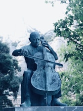 A bronze statue of a person playing a cello is surrounded by lush green trees and a mountainous background. The sculpture captures the intricate details of the musician's posture and the cello.
