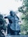 A bronze statue of a person playing a cello is surrounded by lush green trees and a mountainous background. The sculpture captures the intricate details of the musician's posture and the cello.