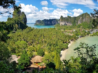 The scene features a tropical landscape with a lush green forest of palm trees leading to a pristine beach. Beyond the beach lies a turquoise sea, flanked by towering, jagged limestone cliffs under a clear blue sky dotted with fluffy white clouds. Thatched-roof huts are nestled among the trees, suggesting a serene, secluded resort setting.