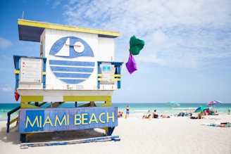 A colorful lifeguard tower displaying a large 'Miami Beach' sign in bold blue and yellow letters stands prominently on the beach. The structure features a unique design with geometric shapes and vibrant colors, adorned with a green and purple flag fluttering atop. The sandy beach spreads out under a partially cloudy sky with turquoise waters visible in the background. Sunbathers and umbrellas dot the landscape, capturing the lively atmosphere of a popular beach destination.