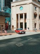 A red taxi is driving on a city street next to a substantial beige building with tall columns and a circular window. Pedestrians are walking on the sidewalk, and the scene is set in an urban environment with other high-rise buildings nearby.