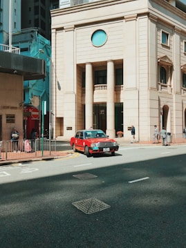 A red taxi is driving on a city street next to a substantial beige building with tall columns and a circular window. Pedestrians are walking on the sidewalk, and the scene is set in an urban environment with other high-rise buildings nearby.