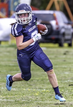 A football player wearing a blue uniform is running with the ball on a grassy field. The player is wearing a helmet with a face guard and padded protection. A parked car and some playground equipment are visible in the background.