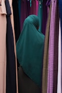 A person wearing a dark green hijab is standing among rows of colorful hanging fabrics. The fabrics include shades of purple, pink, tan, and green, creating a diverse and vibrant backdrop.