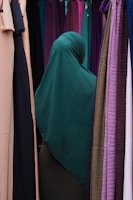 A person wearing a dark green hijab is standing among rows of colorful hanging fabrics. The fabrics include shades of purple, pink, tan, and green, creating a diverse and vibrant backdrop.