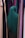 A person wearing a dark green hijab is standing among rows of colorful hanging fabrics. The fabrics include shades of purple, pink, tan, and green, creating a diverse and vibrant backdrop.