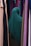 A person wearing a dark green hijab is standing among rows of colorful hanging fabrics. The fabrics include shades of purple, pink, tan, and green, creating a diverse and vibrant backdrop.