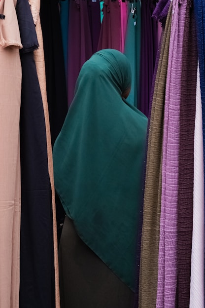 A person wearing a dark green hijab is standing among rows of colorful hanging fabrics. The fabrics include shades of purple, pink, tan, and green, creating a diverse and vibrant backdrop.