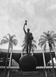 A prominent statue of a figure holding a trophy stands atop a large globe-like base inscribed with 'BRASIL 1958 1962'. The statue is surrounded by tall palm trees, and in the background, there's a large circular building with columns.