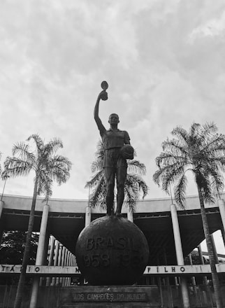 A prominent statue of a figure holding a trophy stands atop a large globe-like base inscribed with 'BRASIL 1958 1962'. The statue is surrounded by tall palm trees, and in the background, there's a large circular building with columns.