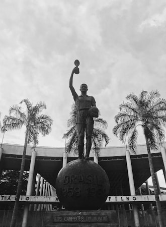 A prominent statue of a figure holding a trophy stands atop a large globe-like base inscribed with 'BRASIL 1958 1962'. The statue is surrounded by tall palm trees, and in the background, there's a large circular building with columns.