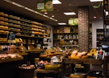 A cozy and well-organized cheese shop filled with an extensive variety of cheeses neatly displayed on shelves. The shop features rustic brick walls, warm lighting, and an assortment of other gourmet products such as bread and preserves. The ambiance is inviting and speaks to a traditional and authentic shopping experience.