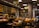 A cozy and well-organized cheese shop filled with an extensive variety of cheeses neatly displayed on shelves. The shop features rustic brick walls, warm lighting, and an assortment of other gourmet products such as bread and preserves. The ambiance is inviting and speaks to a traditional and authentic shopping experience.