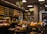 A cozy and well-organized cheese shop filled with an extensive variety of cheeses neatly displayed on shelves. The shop features rustic brick walls, warm lighting, and an assortment of other gourmet products such as bread and preserves. The ambiance is inviting and speaks to a traditional and authentic shopping experience.