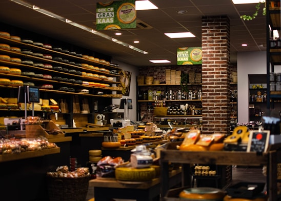A cozy and well-organized cheese shop filled with an extensive variety of cheeses neatly displayed on shelves. The shop features rustic brick walls, warm lighting, and an assortment of other gourmet products such as bread and preserves. The ambiance is inviting and speaks to a traditional and authentic shopping experience.
