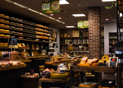A cozy and well-organized cheese shop filled with an extensive variety of cheeses neatly displayed on shelves. The shop features rustic brick walls, warm lighting, and an assortment of other gourmet products such as bread and preserves. The ambiance is inviting and speaks to a traditional and authentic shopping experience.