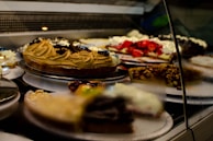 A display case filled with various types of cakes and desserts, including a caramel swirl cake, a strawberry-topped pastry, and a dessert with nuts. The arrangement is behind glass, highlighting the vibrant colors and textures of the treats.
