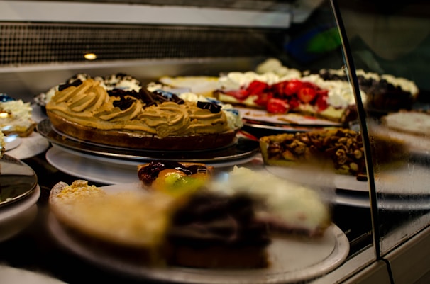 A display case filled with various types of cakes and desserts, including a caramel swirl cake, a strawberry-topped pastry, and a dessert with nuts. The arrangement is behind glass, highlighting the vibrant colors and textures of the treats.