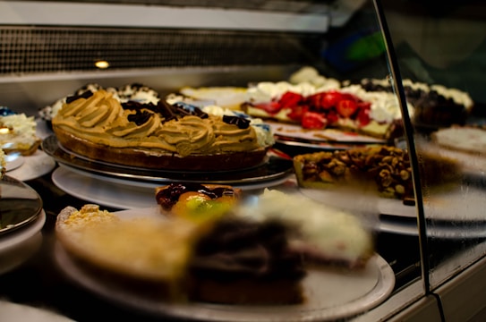 A display case filled with various types of cakes and desserts, including a caramel swirl cake, a strawberry-topped pastry, and a dessert with nuts. The arrangement is behind glass, highlighting the vibrant colors and textures of the treats.