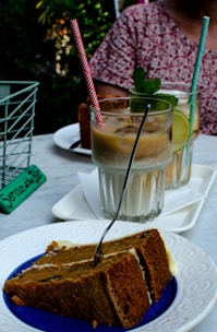 A slice of cake on a white decorative plate with a fork in it, positioned in the foreground. Behind the cake, there are two glasses of iced coffee, each with a colorful straw. The drinks are garnished with mint leaves and lime slices. A person in a patterned shirt is partially visible in the background, and there is a green basket with a service sign on the table.