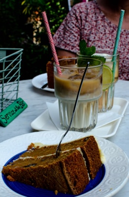 A slice of cake on a white decorative plate with a fork in it, positioned in the foreground. Behind the cake, there are two glasses of iced coffee, each with a colorful straw. The drinks are garnished with mint leaves and lime slices. A person in a patterned shirt is partially visible in the background, and there is a green basket with a service sign on the table.