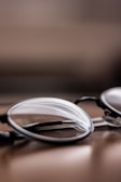 A close-up view of a pair of eyeglasses, positioned on a smooth, reflective surface. The lenses and frames are in focus while the background is blurred, creating a depth of field effect.