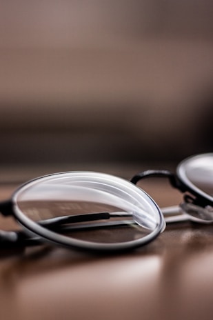A close-up view of a pair of eyeglasses, positioned on a smooth, reflective surface. The lenses and frames are in focus while the background is blurred, creating a depth of field effect.