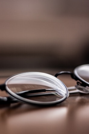A close-up view of a pair of eyeglasses, positioned on a smooth, reflective surface. The lenses and frames are in focus while the background is blurred, creating a depth of field effect.